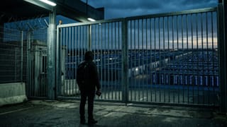 Person facing locked gate before vast server farm at dusk, passport in hand, barbed wire above.