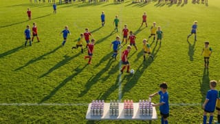 Youth soccer players compete on a sunlit field as Chobani drinks line a sideline table.