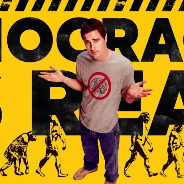 In the image, Luke Wilson, star of Idiocracy, poses on yellow background wearing anti-brain shirt, suggesting devolution.